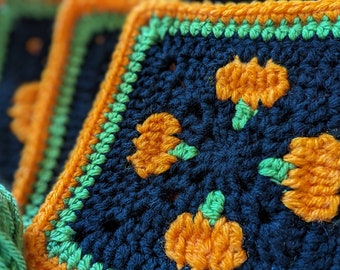 May include: Close-up of a crocheted square with a black background and orange pumpkins. The pumpkins have green stems and are surrounded by a green border.