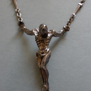 May include: A silver-toned metal crucifix pendant featuring a detailed, textured figure of a person with arms outstretched, suspended from a delicate metal chain with elongated links. The metal has a dark, oxidised finish with visible highlights.