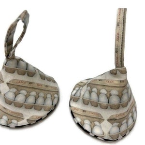 May include: Two brown and white doorstops shaped like a wedge with a loop handle. The doorstops are covered in fabric with a pattern of egg cartons and the words "Large Eggs" and "Eggs".