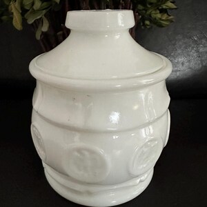 Puede incluir: Un tarro blanco opaco con tapa. El tarro tiene un cuerpo redondeado, una base acampanada y diseños circulares en relieve. Bandas horizontales rodean el tarro. La tapa encaja bien, y el diseño es simple y elegante.