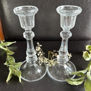 May include: A pair of clear glass candle holders featuring a fluted top, a slender stem with a central decorative ring, and a wide circular base. The candle holders are positioned side by side with small sprigs of dried white flowers nearby.