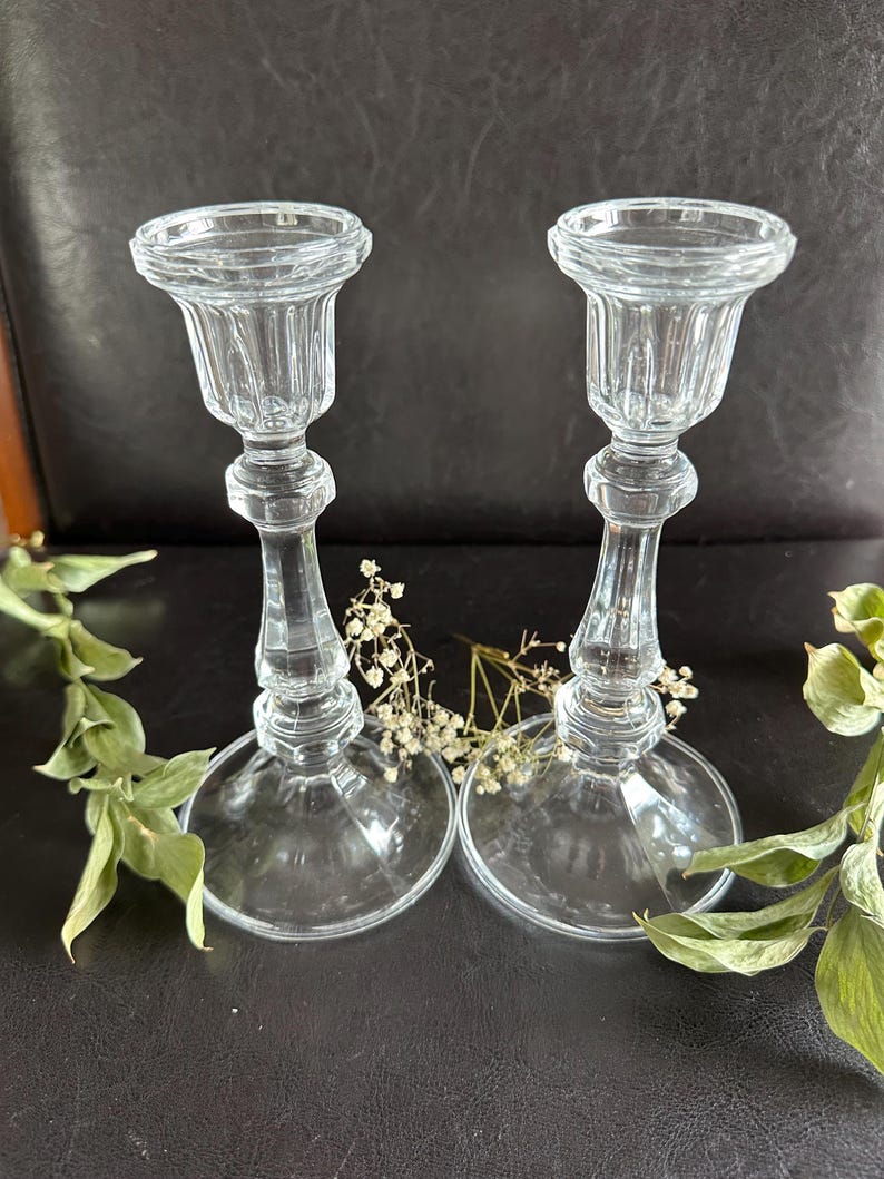 May include: A pair of clear glass candle holders featuring a fluted top, a slender stem with a central decorative ring, and a wide circular base. The candle holders are positioned side by side with small sprigs of dried white flowers nearby.
