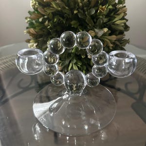 Op de afbeelding: Transparante glazen kandelaar met een cirkelvormig ontwerp van verbonden bollen. De houder heeft twee bekervormige openingen voor kaarsen en een ronde basis. Een groene plant is op de achtergrond.