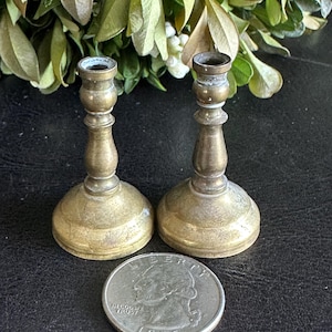 Könnte beinhalten: Zwei kleine, antike Messing-Kerzenhalter mit einer Münze zum Größenvergleich. Die Kerzenhalter haben einen runden Sockel, einen schlanken Stiel und eine kleine Tasse oben. Die Münze ist silberfarben. Die Kerzenhalter sind etwa 7 cm hoch.