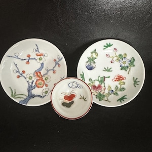 May include: Three small, white ceramic plates with floral designs. Two larger plates feature detailed flower and branch patterns in red, blue, and green. A smaller plate in the center has a red and green design with a red rim. The plates are on a black surface.