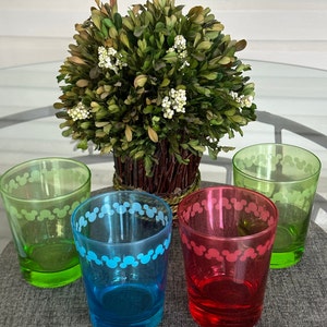 May include: Four glass cups with a Mickey Mouse design around the rim. The cups are green, blue, red, and clear. The cups are sitting on a grey surface in front of a small green bush.