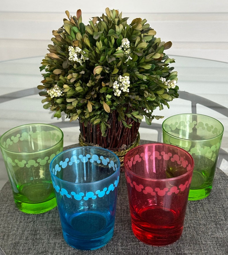 May include: Four glass cups with a Mickey Mouse design around the rim. The cups are green, blue, red, and clear. The cups are sitting on a grey surface in front of a small green bush.