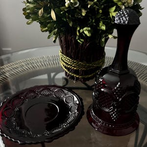 Op de afbeelding: Een donkerrode glazen bel en bijpassende schotel met sierlijke details. De bel heeft een getextureerde bovenkant en een uitlopende basis. De schotel heeft een decoratieve rand. De objecten staan op een glazen tafel met een bloemstuk op de achtergrond.