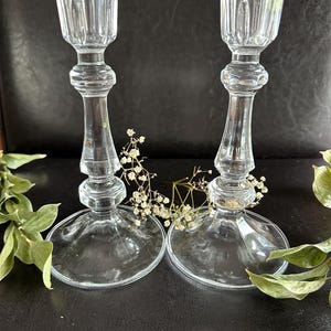 May include: Two clear glass candle holders featuring a fluted top, a slender stem with decorative rounded nodes, and a wide circular base. The candle holders are positioned side by side, with small sprigs of dried white baby's breath flowers.