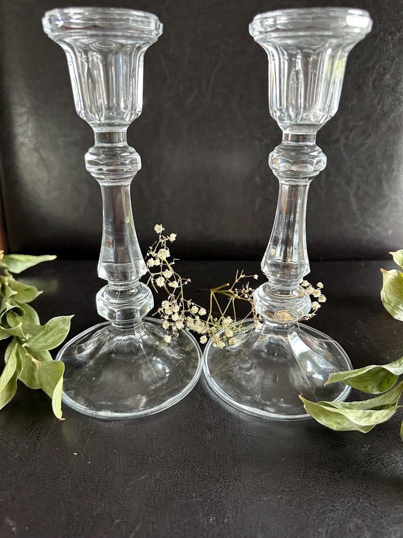 May include: Two clear glass candle holders featuring a fluted top, a slender stem with decorative rounded nodes, and a wide circular base. The candle holders are positioned side by side, with small sprigs of dried white baby's breath flowers.