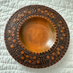 May include: A round wooden bowl with a carved design of leaves and circles. The bowl is brown with a dark brown border.