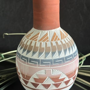 May include: A terracotta vase with a narrow neck and a rounded body featuring carved geometric patterns. The surface displays horizontal bands of white, soft pink, muted blue, and earthy brown tones with intricate etched triangular motifs.