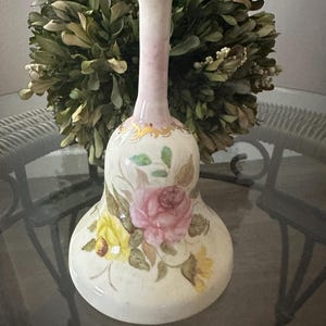 May include: A white ceramic bell with a pink handle and gold accents. The bell is decorated with a floral design featuring pink, yellow, and green flowers.