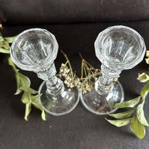 May include: A pair of clear glass candle holders featuring a faceted, multi-sided top rim and a tapered stem leading to a wide, circular base. The glass has a reflective, polished finish with vertical ribbing details along the upper section.