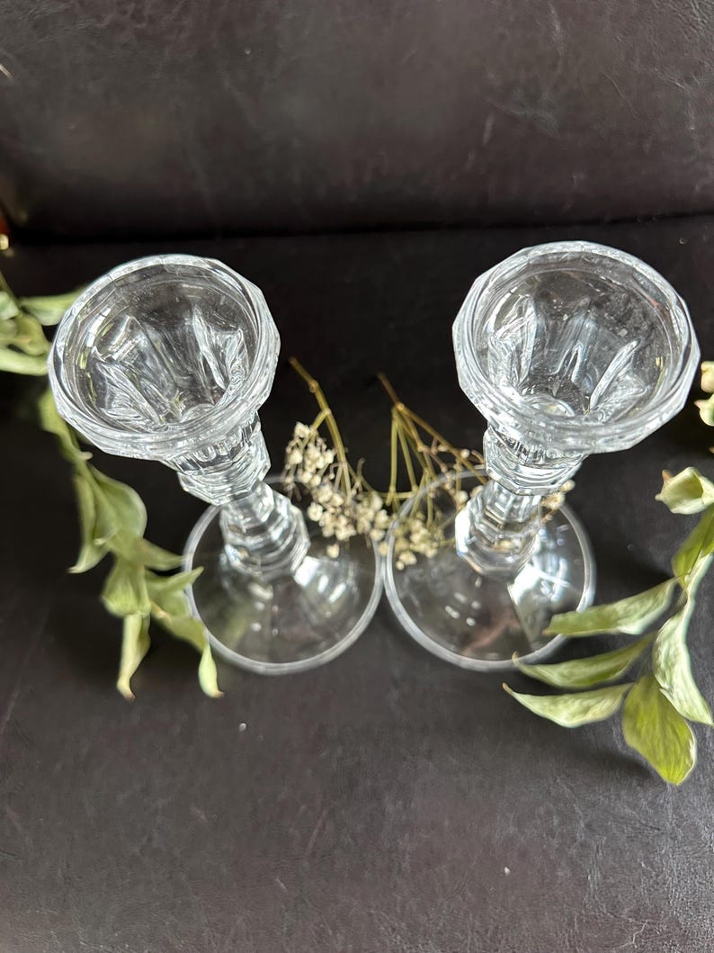 May include: A pair of clear glass candle holders featuring a faceted, multi-sided top rim and a tapered stem leading to a wide, circular base. The glass has a reflective, polished finish with vertical ribbing details along the upper section.