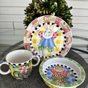 May include: A children's 3-piece dinnerware set featuring a whimsical design. A white plate with a yellow and black checkered border features a white rabbit dressed as a jester. A white bowl with a green and yellow border features a brown bear dressed as a jester. A white mug with a green and yellow border features a monkey dressed as a jester.