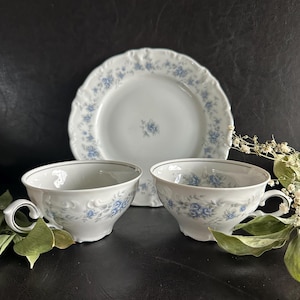 May include: Two white porcelain teacups and a matching saucer featuring a delicate blue floral pattern. The pieces have scalloped edges and a subtle embossed texture along the rims. The design includes small blue rose clusters on the white surface.