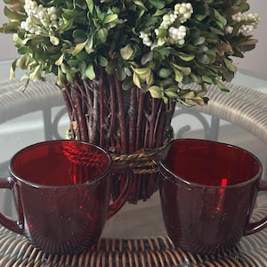 May include: Two ruby red glass mugs with handles are displayed on a glass table. The mugs are in front of a decorative planter filled with greenery and white berries. The mugs have a simple, classic design.