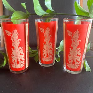 May include: Three clear glass tumblers featuring a vibrant red rectangular panel on the front. Each glass displays a detailed gold-toned graphic of a standing figure in traditional attire holding a staff, accented by vertical gold lines on the sides.