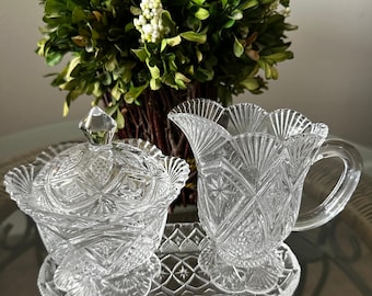 May include: A clear pressed glass creamer and a lidded sugar bowl set resting on a matching rectangular tray. Each piece features a detailed geometric starburst and diamond pattern with scalloped edges, reflecting light through the transparent glass.