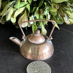 May include: A small, antique copper-colored teapot with a handle and spout. The teapot is placed next to a U.S. quarter coin. Green foliage is in the background. The teapot is likely a decorative item or part of a miniature collection.