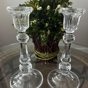 May include: A pair of clear glass candle holders featuring a fluted top, a slender stem with decorative faceted nodes, and a wide circular base. The transparent glass construction reflects light, showcasing a classic vertical design profile.