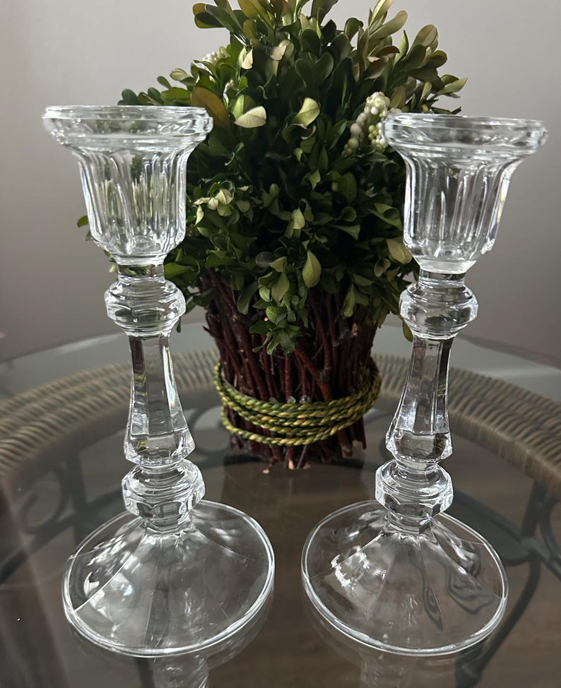 May include: A pair of clear glass candle holders featuring a fluted top, a slender stem with decorative faceted nodes, and a wide circular base. The transparent glass construction reflects light, showcasing a classic vertical design profile.