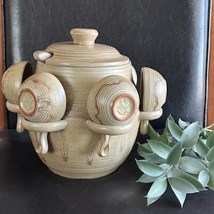 May include: A beige ceramic soup tureen with a lid and four attached soup bowls. The tureen has a round shape with a textured surface and a small handle on the lid. The bowls are round with a handle and a red rim.