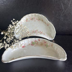 May include: Two antique, off-white, crescent-shaped dishes with floral patterns in pink, green, and gold. The edges are trimmed with gold. The dishes are displayed on a dark surface.