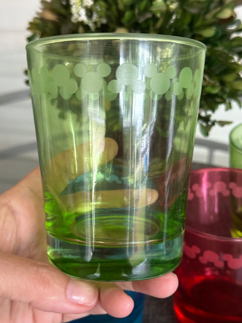 May include: A green glass drinking glass with a white Mickey Mouse design around the top edge.