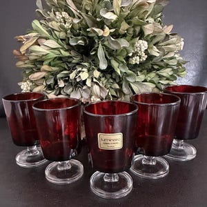 Puede incluir: Juego de seis copas de vino vintage de color rojo rubí. Las copas tienen una base clara y circular y un tallo corto. Las copas tienen forma cilíndrica con un borde ligeramente ensanchado. Una etiqueta es visible en una copa. Un arreglo floral decorativo verde y blanco está en el fondo.