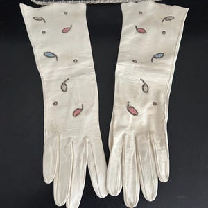 May include: A pair of off-white leather gloves with decorative paisley-shaped accents in pink, blue, and black. The gloves are long, extending past the wrist, and are displayed on a dark surface. A clear glass dish is in the background.