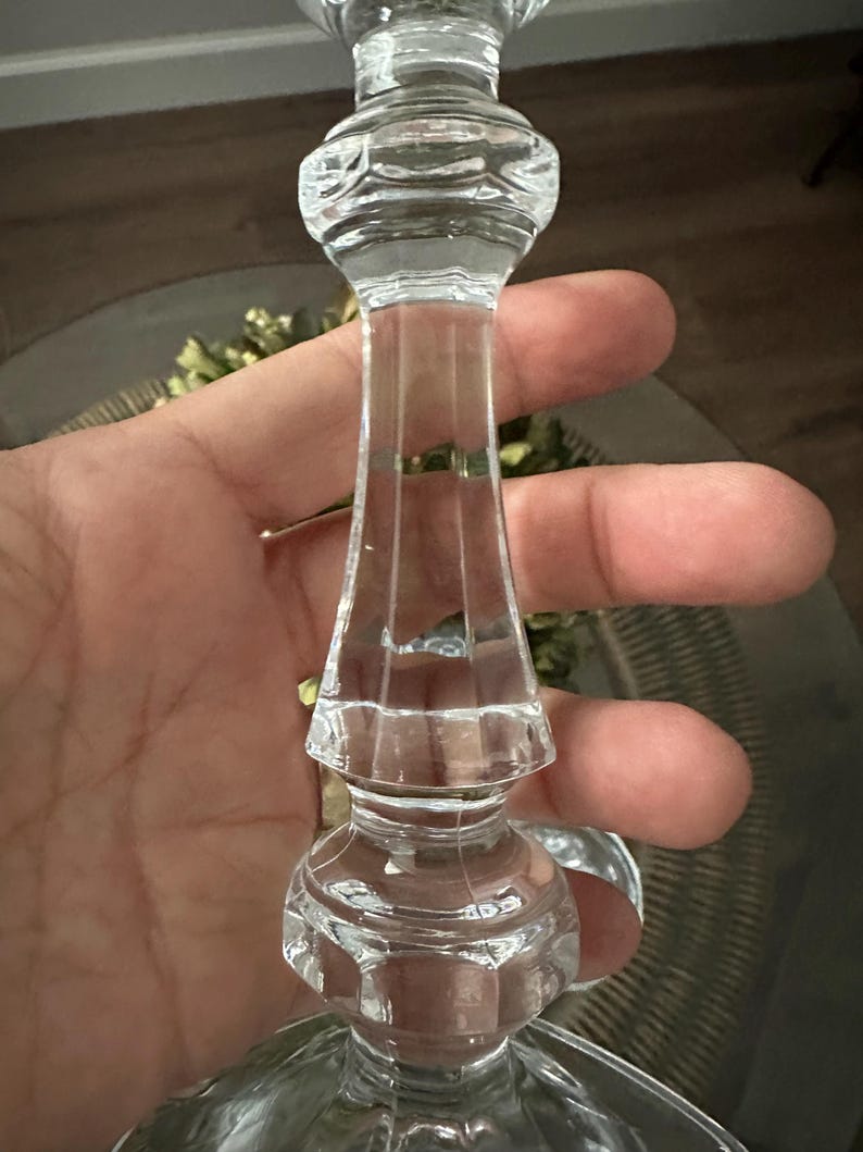 May include: A clear glass candlestick holder featuring a multi-faceted stem with a series of stacked, rounded, and geometric decorative nodes. The glass is transparent and smooth, showcasing a vertical panel design along the central shaft.