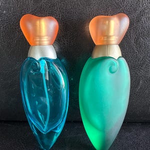May include: Two teardrop-shaped perfume bottles with orange heart-shaped caps and silver collars. The left bottle is made of clear blue glass, while the right bottle is made of frosted green glass. Both feature a swirling embossed design on the front.