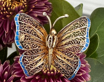 May include: A gold-toned butterfly brooch featuring intricate filigree metalwork on the wings and body. The outer edges of the wings are accented with bright blue enamel detailing. Two thin antennae with small rounded tips extend upward.
