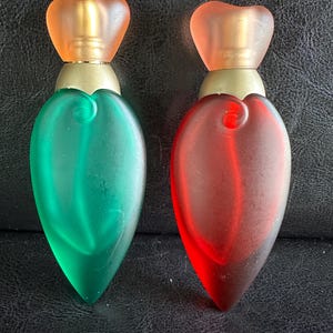 Peut inclure: Deux flacons de parfum décoratifs. L'un est vert, l'autre rouge. Les deux ont un bouchon orange en forme de cœur et un col doré. Les flacons sont en forme de goutte et reposent sur une surface sombre.