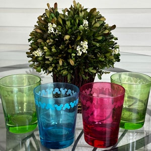 May include: Four glass tumblers with Mickey Mouse silhouettes printed on them. The tumblers are green, blue, red, and clear. They are arranged on a table with a green bush in the background.