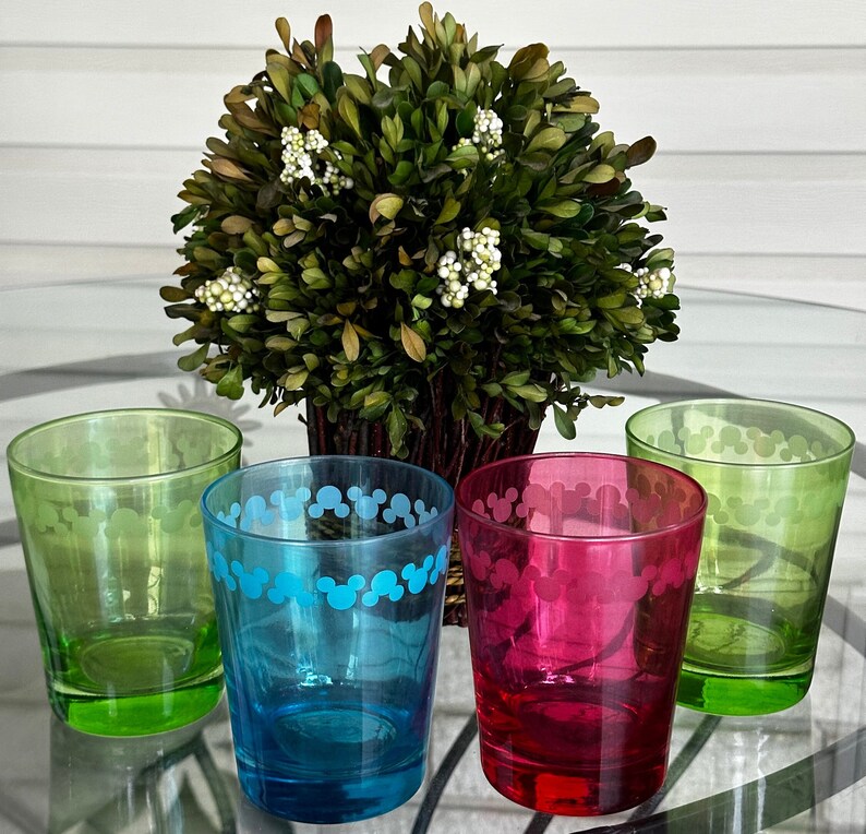 May include: Four glass tumblers with Mickey Mouse silhouettes printed on them. The tumblers are green, blue, red, and clear. They are arranged on a table with a green bush in the background.