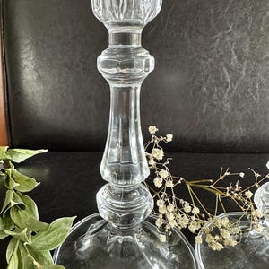 May include: A clear glass candlestick holder featuring a tall, slender stem with a multi-faceted, ribbed design and a wide, circular base. The top section has a vertical fluted pattern, and the central stem displays a series of stacked, rounded nodes.