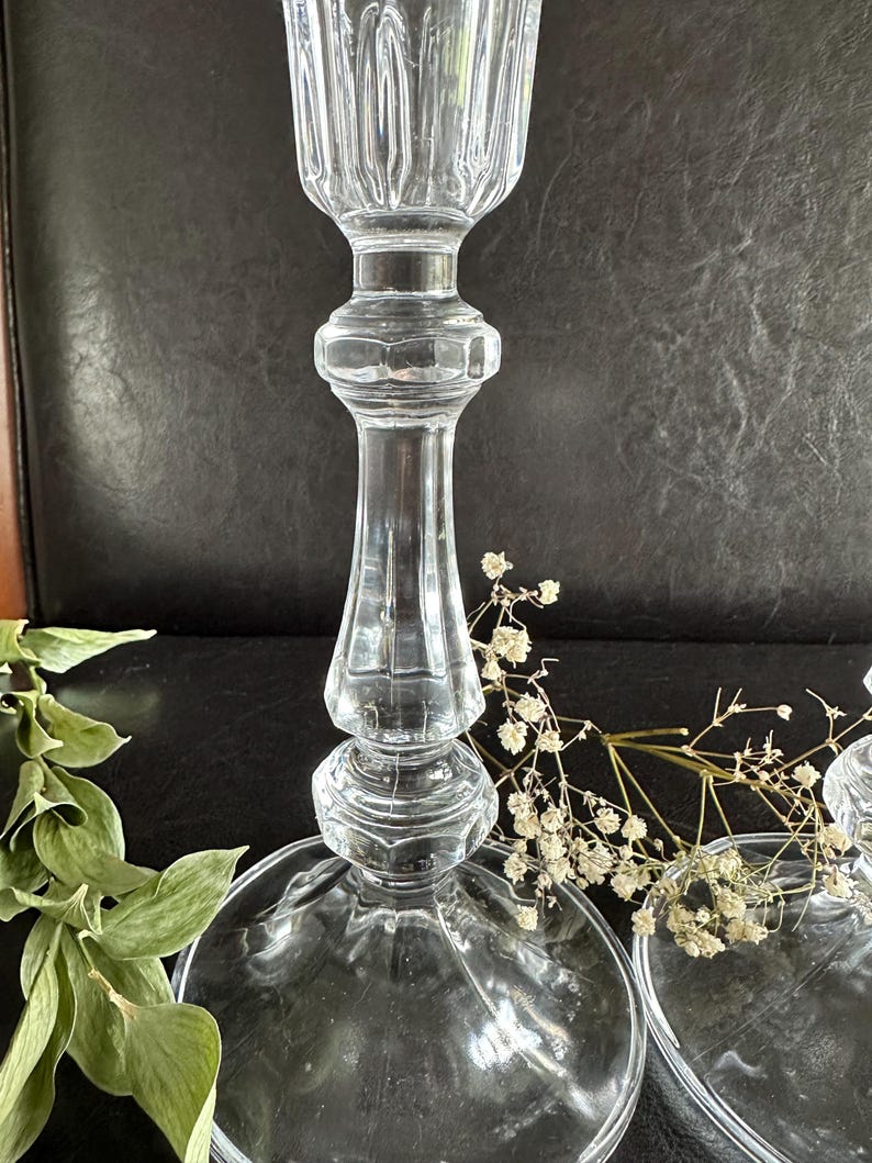 May include: A clear glass candlestick holder featuring a tall, slender stem with a multi-faceted, ribbed design and a wide, circular base. The top section has a vertical fluted pattern, and the central stem displays a series of stacked, rounded nodes.