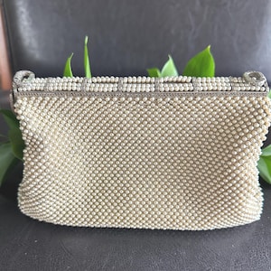 May include: A rectangular clutch bag completely covered in small, off-white faux pearls arranged in a grid pattern. The top frame is made of silver-toned metal, featuring a row of pearls and intricate, textured detailing along the edges.