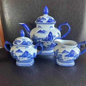 May include: A blue and white porcelain tea set featuring a teapot, sugar bowl, and creamer. The set has a landscape design with mountains and trees. The teapot has a lid with a blue finial, and the handles and rims are also blue.