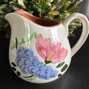 Puede incluir: Una jarra de cerámica de color crema con un diseño pintado a mano de uvas azules, hojas verdes y una flor rosa grande. El interior de la jarra es de color naranja terracota e incluye un asa curva y un pico vertedor para servir líquidos.