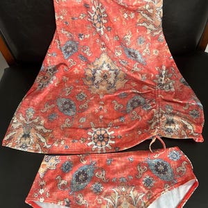 Puede incluir: Un conjunto de traje de baño de dos piezas con top halter y braguita de cintura alta. El conjunto está estampado con un diseño rojo, azul y beige que recuerda a una alfombra vintage. El top tiene un detalle fruncido lateral y la braguita tiene forro blanco.