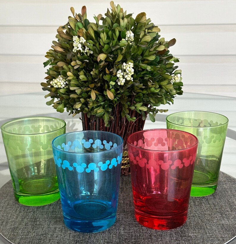 May include: Four clear glass tumblers with a Mickey Mouse design. The tumblers are green, blue, red, and clear. The tumblers are sitting on a table with a green plant in the background.