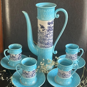 May include: A tall, light blue ceramic coffee pot with a curved handle and spout, paired with four matching cups and saucers. The set features a textured, woven-like pattern and a central band decorated with a detailed blue and white landscape scene.