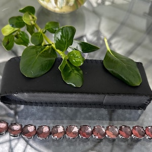 Op de afbeelding: Een zilveren armband met ovale, roze edelstenen. De armband wordt gepresenteerd op een zwarte leren etui, met groene bladeren en een glazen kom op de achtergrond. De edelstenen zijn gezet in een zilveren metalen zetting.