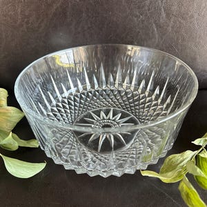 May include: A clear glass bowl featuring a faceted diamond pattern on the exterior and a starburst design at the center of the base. The bowl has a circular rim and a thick, textured wall construction with the word Arcoroc embossed on the bottom.
