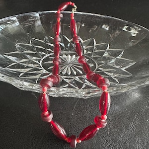 Könnte beinhalten: Eine Vintage-Halskette mit tiefroten Glasperlen in verschiedenen Formen. Die Perlen sind mit einem goldfarbenen Verschluss aufgefädelt. Die Halskette wird auf einer durchsichtigen, geschliffenen Glasschale präsentiert.