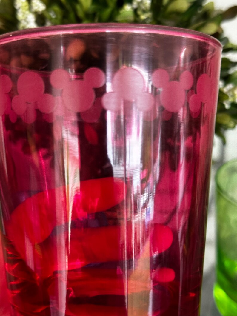 May include: A pink glass with a Mickey Mouse design. The glass is filled with a red liquid.